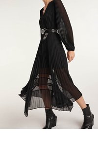 Frau, die ein schwarzes, plissee Midi-Kleid mit durchsichtigen Ärmeln und Saum trägt, einen breiten schwarzen Gürtel mit silbernen Details und schwarze Knöchelstiefel.