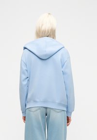 Personne aux cheveux blond platine, portant un sweat à capuche bleu clair et un jean ample en denim clair, debout de dos devant un fond uni.