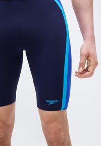 Κάτω μέρος κορμού ατόμου που φοράει μπλε σκούρο Speedo μαγιό τύπου jammers με ανοιχτό και σκούρο μπλε ρίγες στα πλευρά, σε λευκό φόντο.