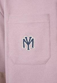 Tasca in tessuto lilla strutturato su un indumento con lettere NY ricamate sovrapposte blu navy e bianche, centrate sulla tasca.