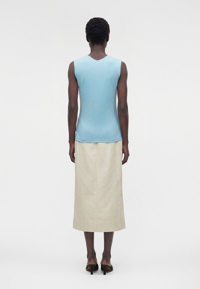 Άτομο με κοντά σγουρά μαλλιά που φοράει γαλάζιο χωρίς μανίκια τοπ, μπεζ midi φούστα και μαύρα σανδάλια με τακούνι, στέκεται γυρισμένο προς τα πίσω σε απλό φόντο.