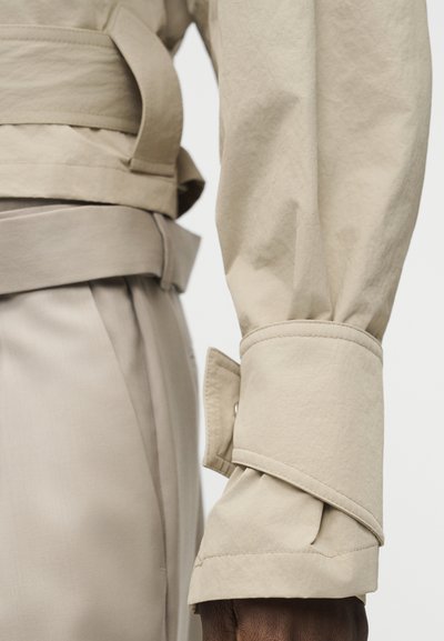 Μπεζ πουκάμισο με μακριά μανίκια και φαρδύ μανσέτα και μπεζ παντελόνια με ζώνη, με κοντινή λήψη των λεπτομερειών της μέσης και του μπράτσου.