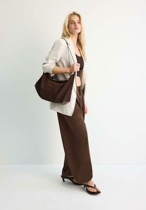 Blond žena nosící bílý blejzr, hnědý kratší top, široké hnědé kalhoty, černé sandály na podpatku, nesoucí velkou hnědou kabelku přes rameno, stojící u bílé zdi.