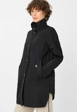 Frau mit kurzen lockigen Haaren, die einen schwarzen, geknöpften Mantel mit hohem Kragen und beigen Hosen trägt und mit den Händen in den Taschen steht.