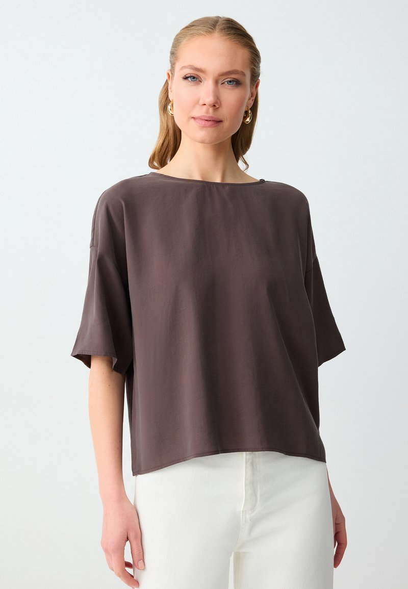 Femme aux cheveux châtain clair portant des boucles d'oreilles créoles en or, un haut ample à manches courtes marron foncé, et un pantalon blanc sur un fond blanc uni.