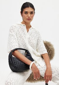 Svart vävd handväska med rund form och flätad handtag, tillsammans med en vit skjorta med spetsstruktur och matchande byxor.