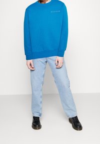 Sudadera azul con cuello redondo, con texto en gris "HILFIGER", combinada con jeans sueltos de azul claro y botas negras con cordones.