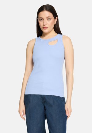 Femme portant un haut sans manches bleu clair avec une petite découpe près du col et un pantalon taille haute bleu foncé, debout devant un fond uni.