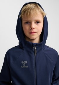 Marinblå huvjacka tillverkad av mjukt tyg; har dragkedja, logotyp på bröstet och ljusgrå accenter på axlarna.