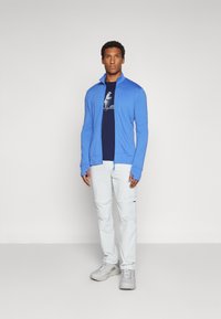 Chaqueta azul con cremallera sobre una camiseta navy, combinada con pantalones blancos desmontables y zapatillas grises. El material parece ligero y suave.