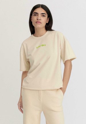 Beige t-shirt met korte mouwen met groene "LOG OFF" tekst. Zacht materiaal, relaxte fit en onafgewerkte zoomdetails. Gecombineerd met lichte onderkleding.