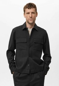 Moški nosi črno jakno s gumbi in dvema žepoma na prsih, preko svetlo sive srajce, stoji z rokama v žepih ob beli ozadju.