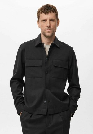 Hombre vestido con una chaqueta negra abotonada con dos bolsillos en el pecho sobre una camisa gris claro, de pie con las manos en los bolsillos contra un fondo blanco.