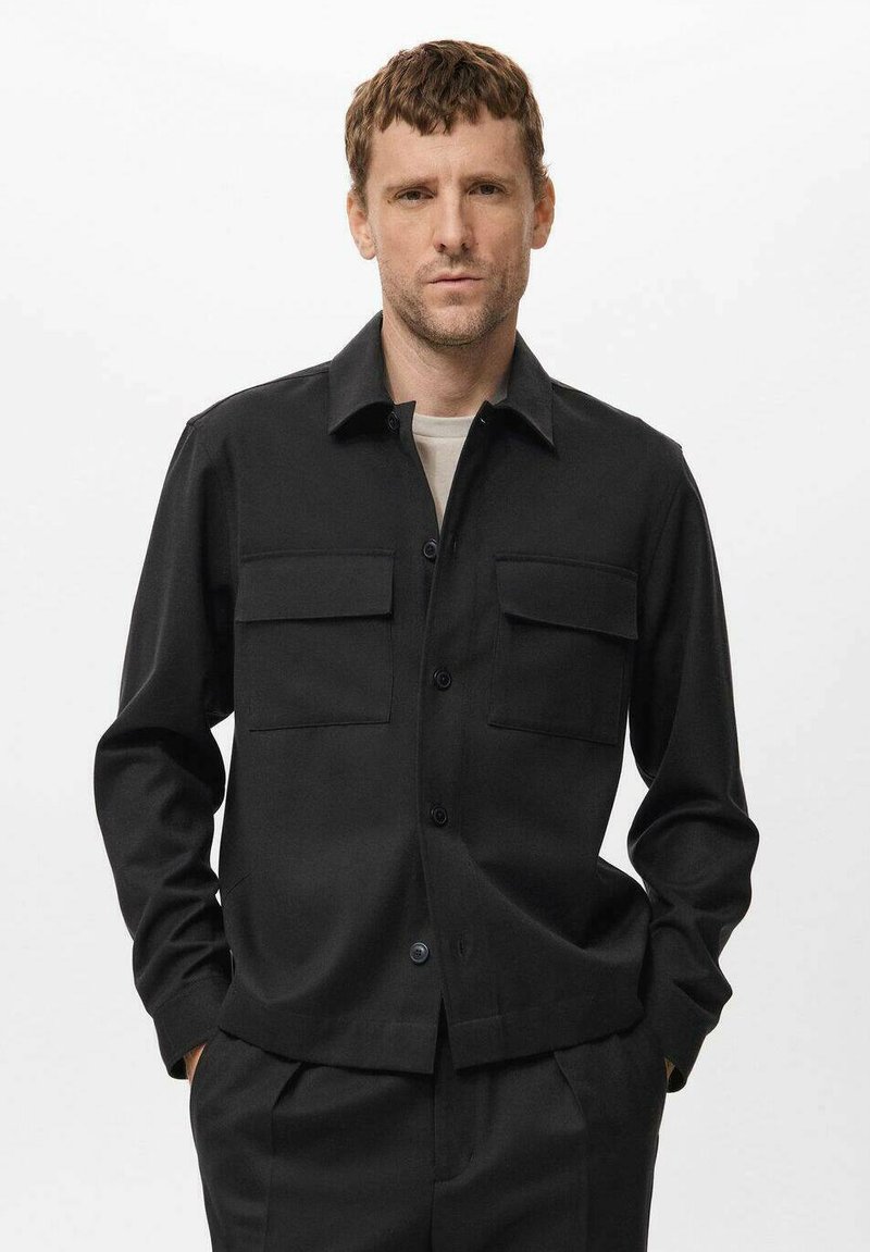 Moški nosi črno jakno s gumbi in dvema žepoma na prsih, preko svetlo sive srajce, stoji z rokama v žepih ob beli ozadju.