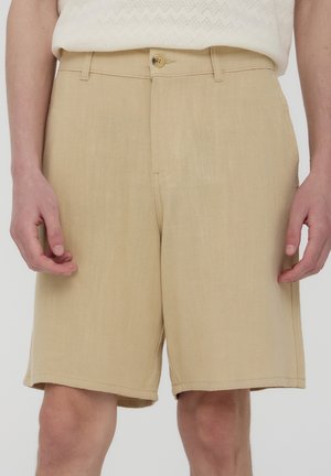 Pantaloni corti beige su misura realizzati in un tessuto testurizzato, con chiusura frontale a bottone e tasche laterali, dal taglio dritto.
