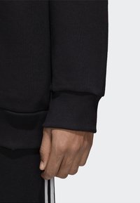 Manga de sudadera negra con textura acanalada, con un puño ajustado y una mano apoyada en la costura, combinada con pantalones negros y franjas blancas.