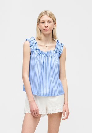 Νεαρή γυναίκα που φοράει μπλε και λευκή ριγέ αμάνικη μπλούζα με βολάν στους ώμους και λευκά σορτς με δαντελένιο τελείωμα, στέκεται μπροστά σε ένα απλό φόντο.