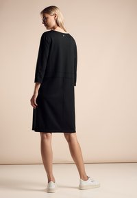 Abito nero di media lunghezza con maniche a tre quarti, scollatura rotonda e dettagli delle cuciture; indossato con sneaker bianche. Tessuto liscio, design casual.