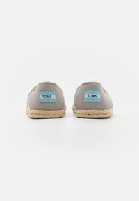 Espadrilles en toile gris avec semelles en jute, présentant une étiquette TOMS bleue à l'arrière et un design classique à enfiler.