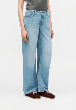 Άτομο που φοράει ανοιχτό μπλε τζιν με φαρδύ πόδι, γκρι αμάνικο τοπ και καφέ παπούτσια με κορδόνια, στέκεται μπροστά σε απλό φόντο.