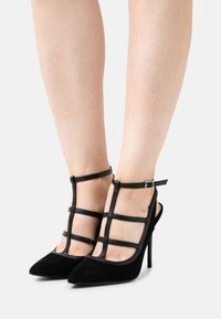 Sapatos altos de camurça pretos com parte traseira aberta, design de tiras, bico apontado e tira de tornozelo com fivela. Textura suave e silhueta elegante.