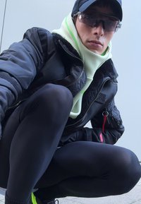 Zwarte bomberjack met bontvoering, limoen groene hoodie, zwarte leggings, donkere sportzonnebril en zwarte pet, zittende pose op een vlakke ondergrond.
