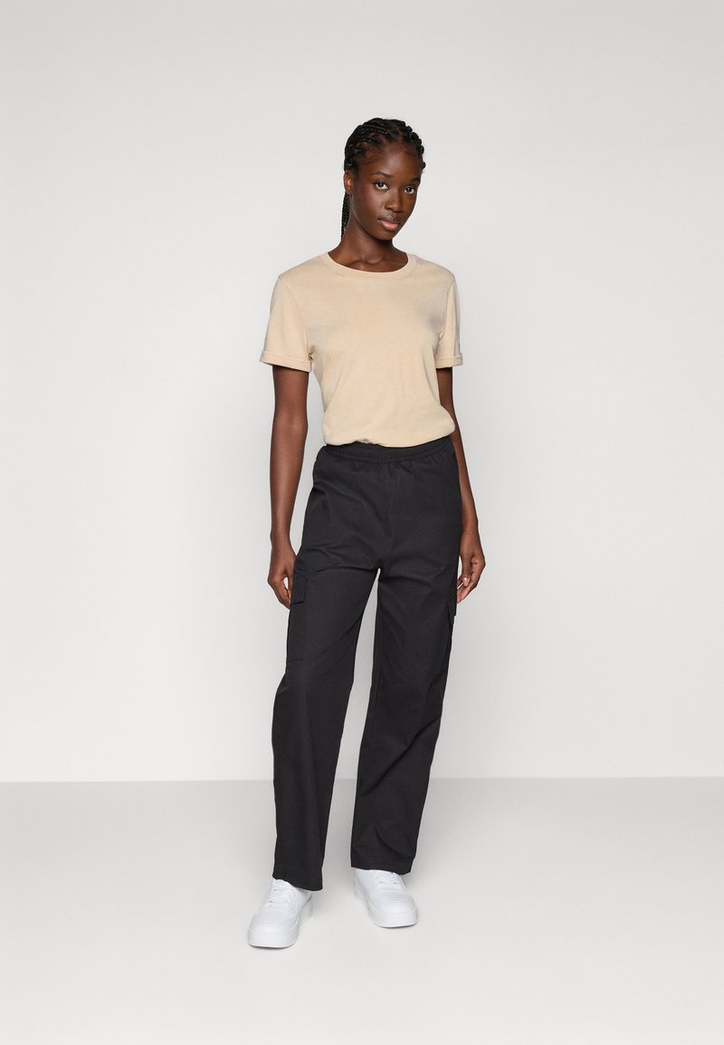 Beige Baumwoll-T-Shirt mit kurzen Ärmeln, kombiniert mit schwarzen Cargo-Hosen mit elastischem Bund und seitlichen Taschen, abgerundet mit weißen Sneakers.