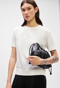 Top blanco de manga corta con cuello redondo, que presenta una textura suave. Bolso negro con pliegues sostenido en una mano, mostrando su material blando.