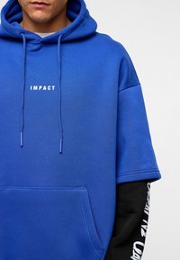 Blauwe hoodie met korte mouwen, met het woord "IMPACT" in witte tekst. Inclusief een voorzak en contrasterende zwarte mouwdetails.