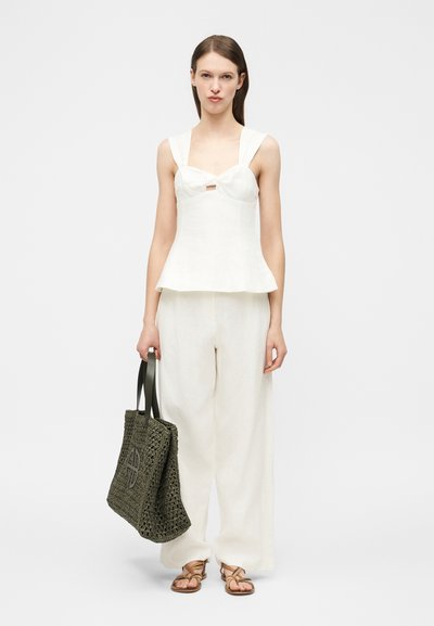 Woman wearing sleeveless white top and wide-leg white pants, holding large black woven tote bag, standing against plain background.