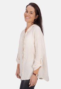 Helle beige, strukturierte Kapuzenbluse mit durchgehender Knopfleiste und hochgerollten Ärmeln. Über schwarzen Leggings getragen.