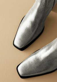 Botas de tobillo metálicas plateadas con un acabado texturizado, punta cuadrada y suelas negras de contraste. Diseñadas con una silueta elegante.