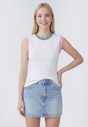 Ung kvinde iført en hvid ærmeløs top med pink og turkis kant og en lyseblå denim nederdel, stående med hånden på hoften og smilende.