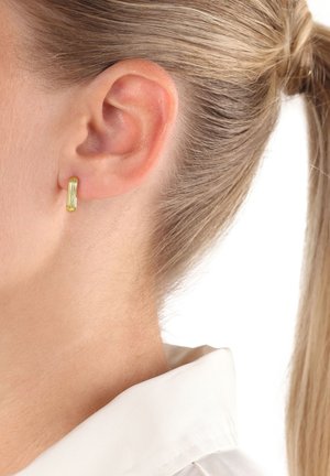 Primer plano de la oreja de una mujer con un pequeño arete de oro en forma de aro, el cabello rubio recogido y una camisa blanca con collar visible.