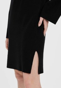 Svart stickad klänning med en sidoslits, som har en mjuk textur och långa ärmar. Slitsen framhäver rörelse och tillför detalj till designen.