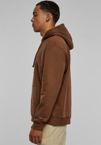 Felpa con cappuccio marrone in una vestibilità comoda, dotata di tasca frontale e polsini a costine. Texture morbida con un colore uniforme e dettagli minimi.