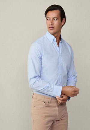 Chemise boutonnée à carreaux bleu clair avec col, en coton, à manches longues et avec un logo subtil, associée à un pantalon beige.