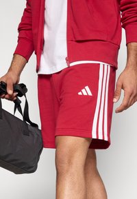 Raudoni sportiniai šortai su trimis baltomis juostelėmis šone, pagaminti iš minkštos medžiagos. Dėvintysis laiko juodą rankinę su dviem rankenomis.