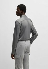 Camicia grigia a maniche lunghe con una texture liscia, colletto a svolazzare e polsi abbottonati, abbinata a pantaloni cargo su misura grigi chiari.