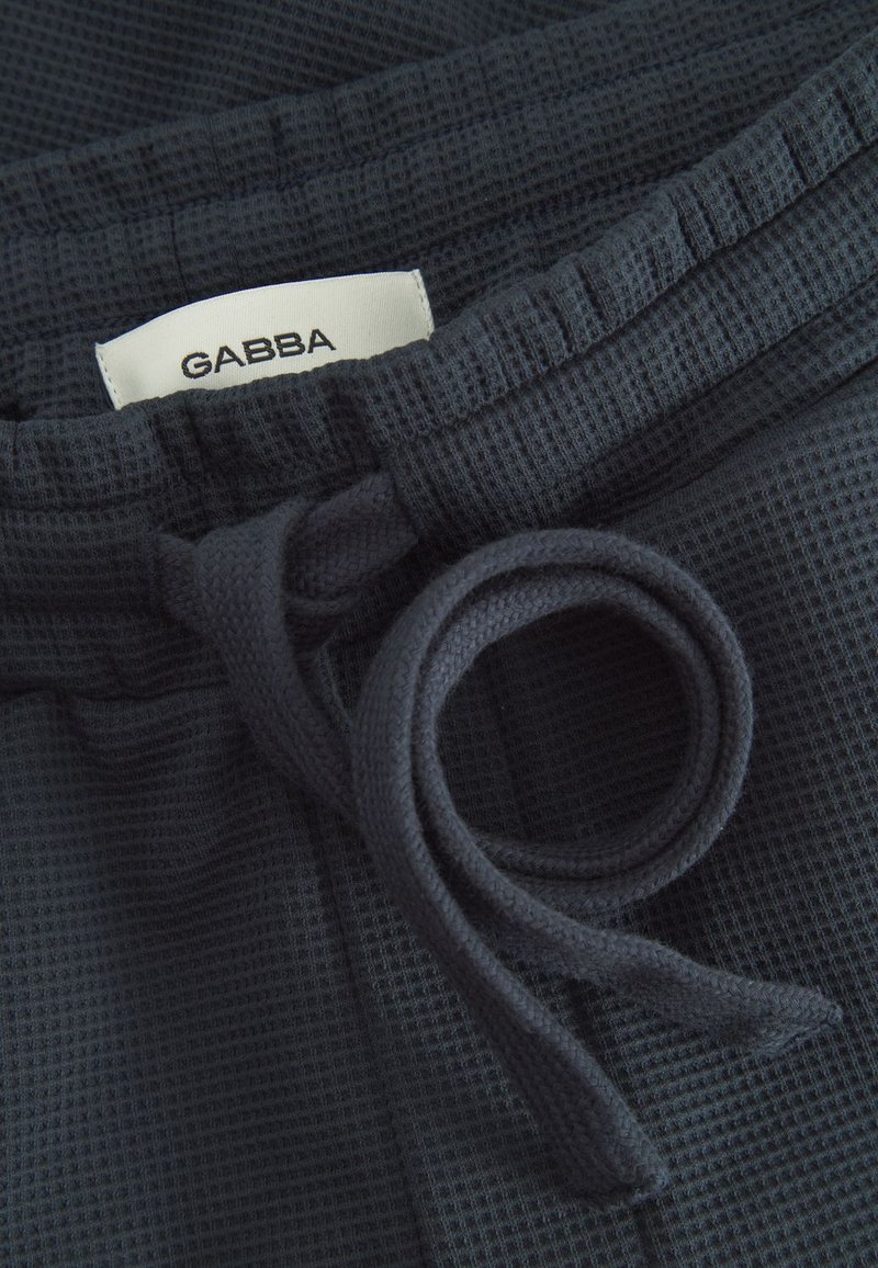 Tumedat merevärvi tekstuuriga vabaajajalanõud, millel on kootud vöökoht, nöör ja silt, millel kiri "GABBA" tikitud märgil.