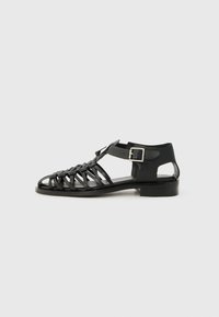 Sandalia de cuero negro con un diseño trenzado, punta abierta y correa en el tobillo. Presenta una hebilla plateada y tacón bajo. Suela plana.