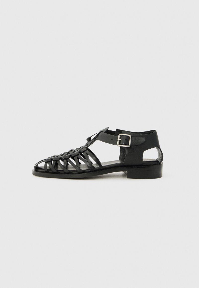 Black leather sandal with a woven design, open toe, and ankle strap. Features a silver buckle and low block heel. Flat sole.