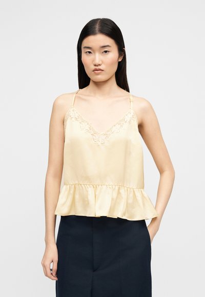 Jeune femme aux cheveux bruns et raides portant un chemisier sans manches jaune pâle orné de dentelle et un pantalon noir taille haute, debout avec une main dans la poche.