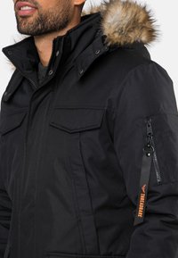 Sort parka-jakke med pelsforet hætte, der har klaplommer, lynlåsdetaillering og et orange mærke. Tekstureret stof og struktureret form.