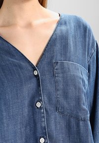 Gros plan sur une personne portant une chemise en jean bleue avec des boutons blancs et une poche poitrine, avec un col en V profond.