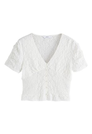 Frau trägt eine strukturierte weiße Kurzarm-Bluse mit Perlenknöpfen und beige hoch taillierte Hose, posiert vor neutralem Hintergrund.