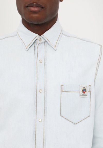 Camicia in denim azzurro chiaro con colletto a punta, tasche frontali e cuciture in tono contrastante color beige. Presenta un'etichetta sulla tasca con il marchio "BALLY".