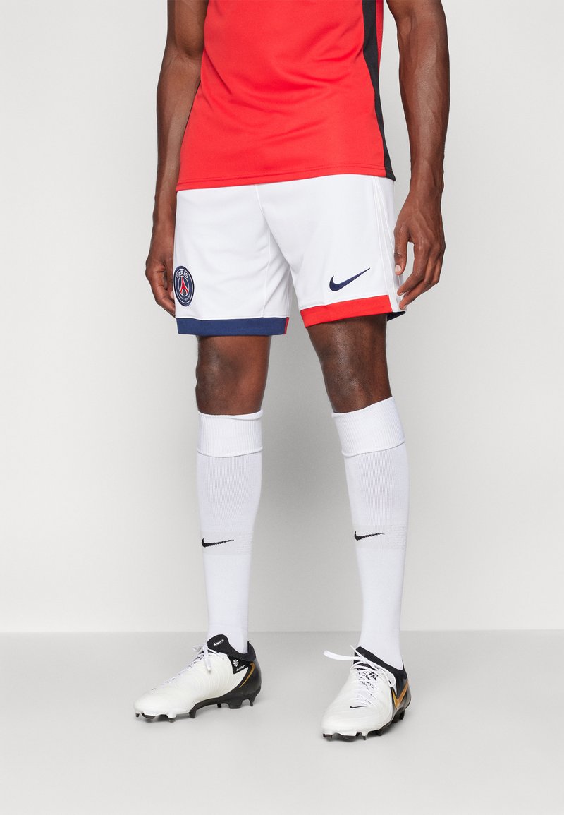 Witte voetbalkortingen met blauwe en rode accenten, voorzien van een logo van Paris Saint-Germain. Gecombineerd met witte sokken en zwart-witte voetbalschoenen.