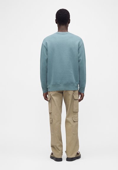 Personne portant un sweat-shirt bleu clair et un pantalon cargo beige, debout de dos sur un fond blanc uni.