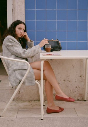 Femme aux cheveux bouclés portant un blazer à carreaux, assise à une table de café en plein air avec une boisson, un sac noir, un magazine, et des ballerines rouges.